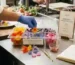 A professional chef arranging edible flowers from a clear multi-compartment container on a stainless steel counter at topofficialgenetics.